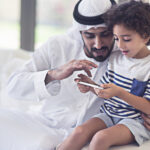 Middle Eastern father sharing time with his son at home, father is teaching how to use a mobile phone application,the father is wearing the traditional Dishdasha.