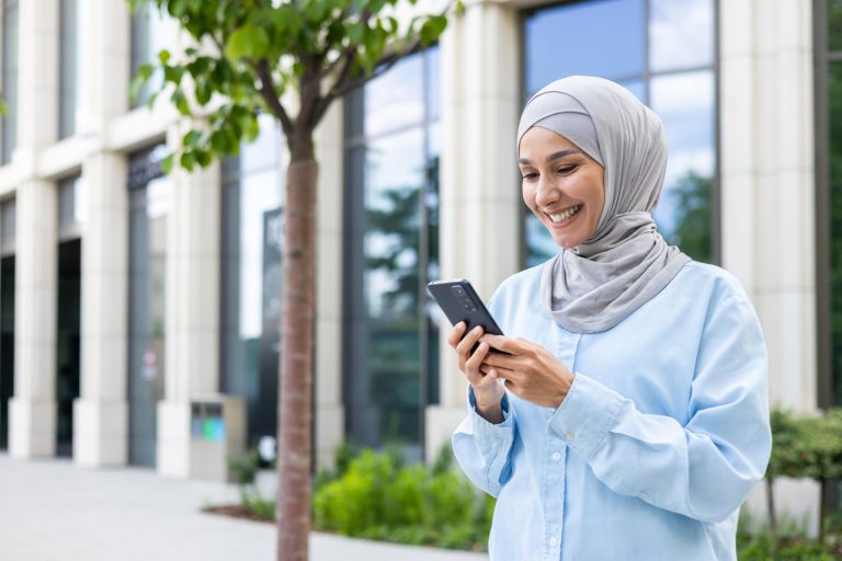 Young beautiful business woman in hijab walks in the city outside the office building, Muslim woman uses an application on the phone, browses online internet pages, smiles contentedly